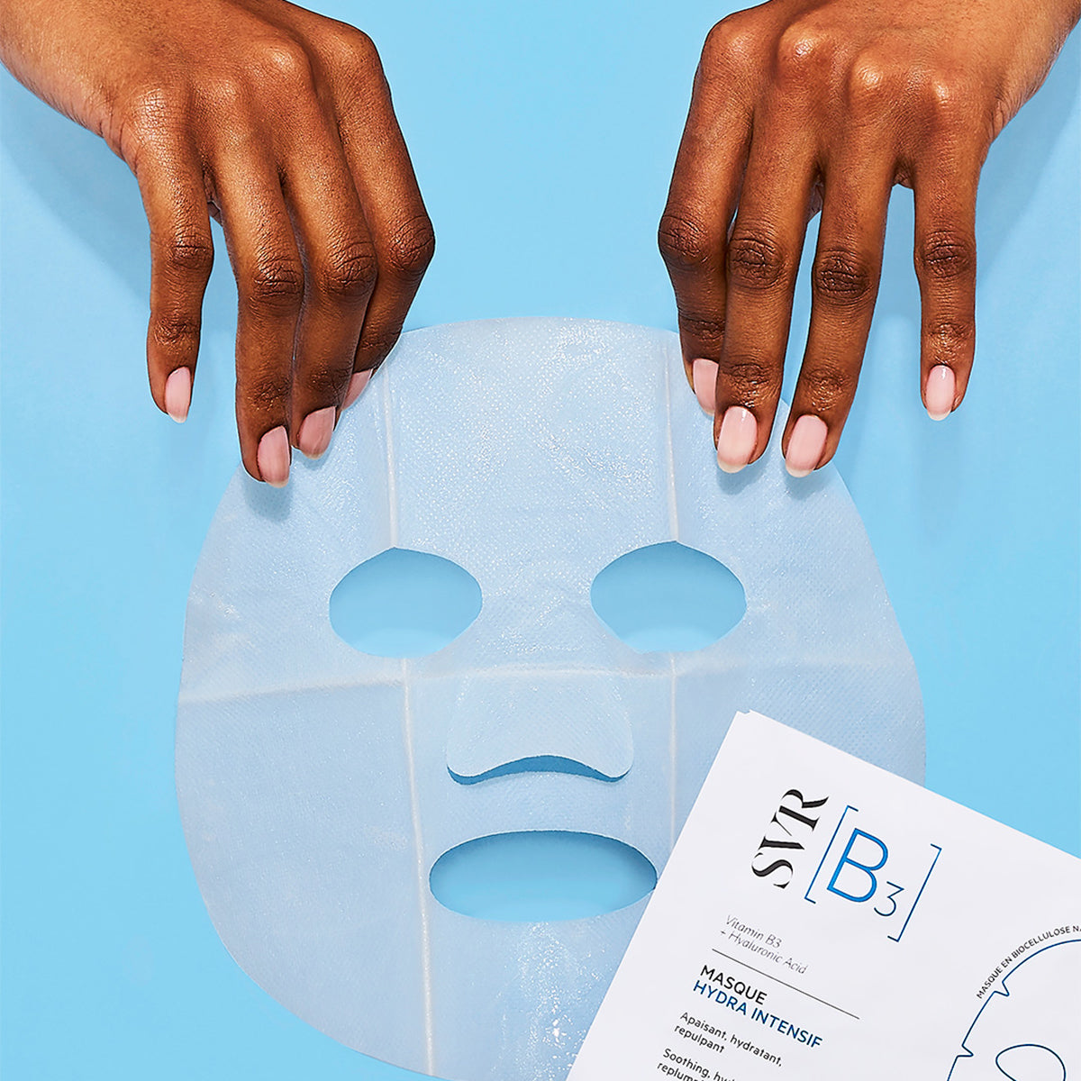 Hands holding a translucent biocellulose SVR B3 hydrating face mask against a light blue background.