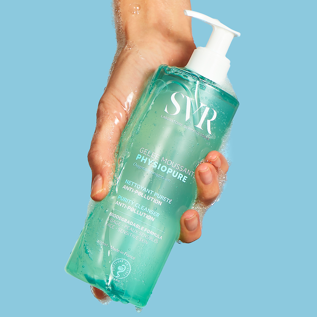 Frasco de gel de limpeza SVR Physiopure com doseador, segurado por uma mão com espuma sobre fundo azul claro.