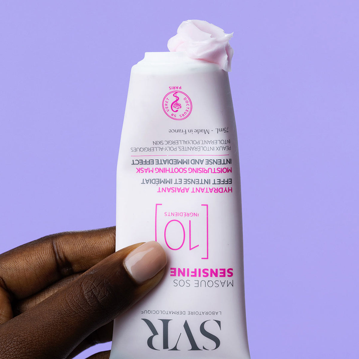 Tubo branco de SVR Sensifine Masque SOS com textura cremosa rosada, segurado por uma mão sobre fundo lilás.