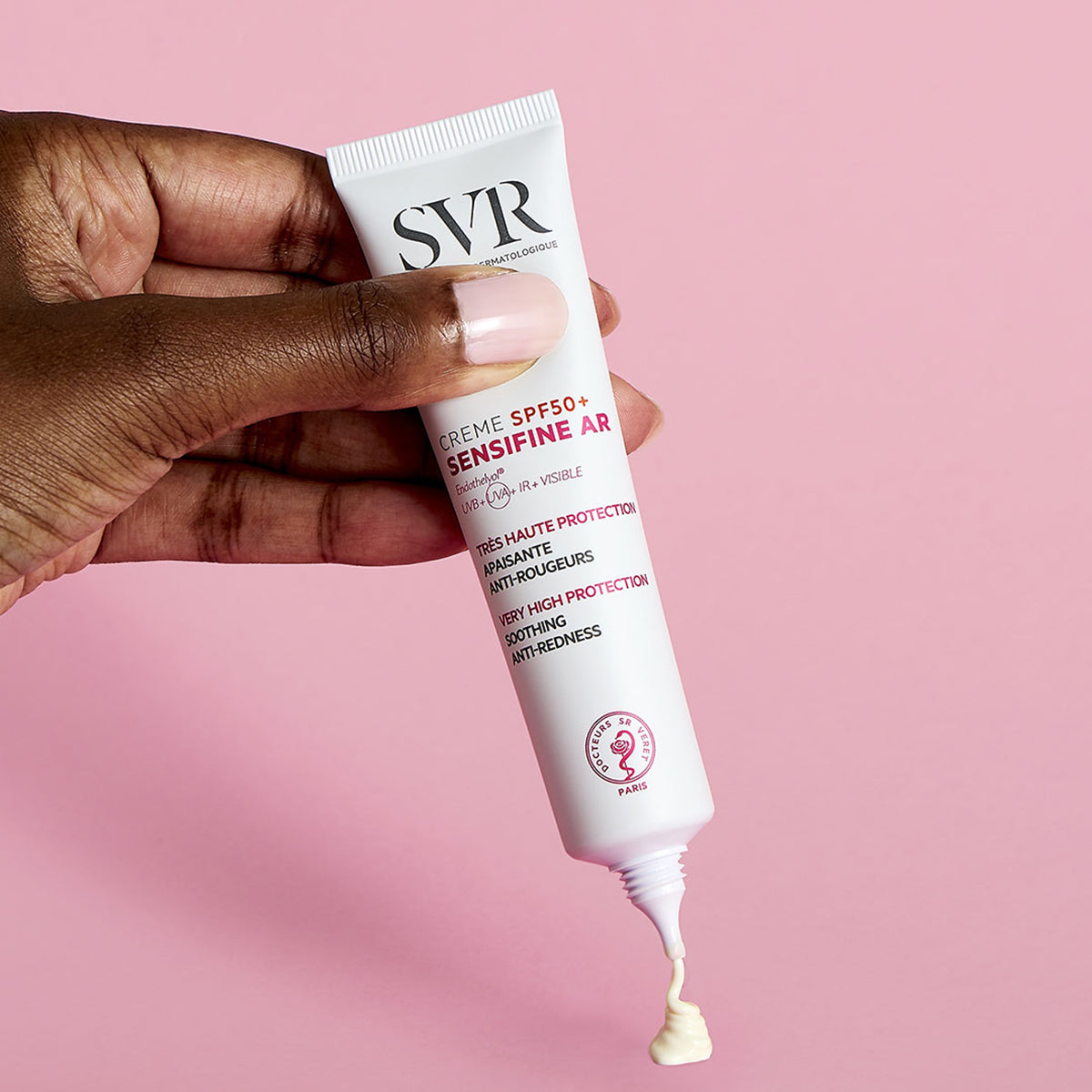 Mão segurando bisnaga branca de SVR Sensifine AR Creme SPF50+ com textura cremosa saindo contra fundo rosa.
