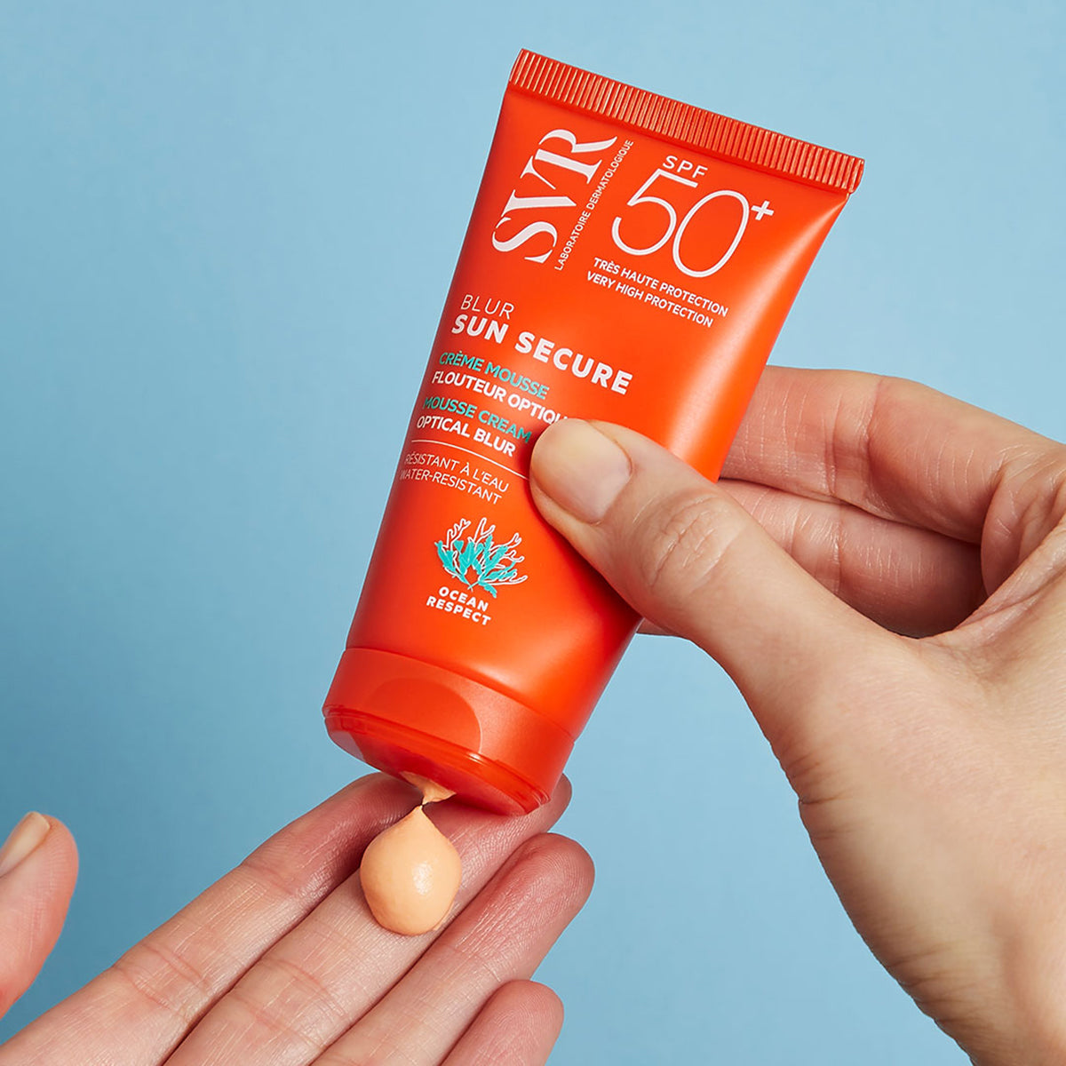 Bisnaga laranja de protetor solar SVR Sun Secure Blur SPF50+ sendo aplicada na palma da mão com textura em mousse.