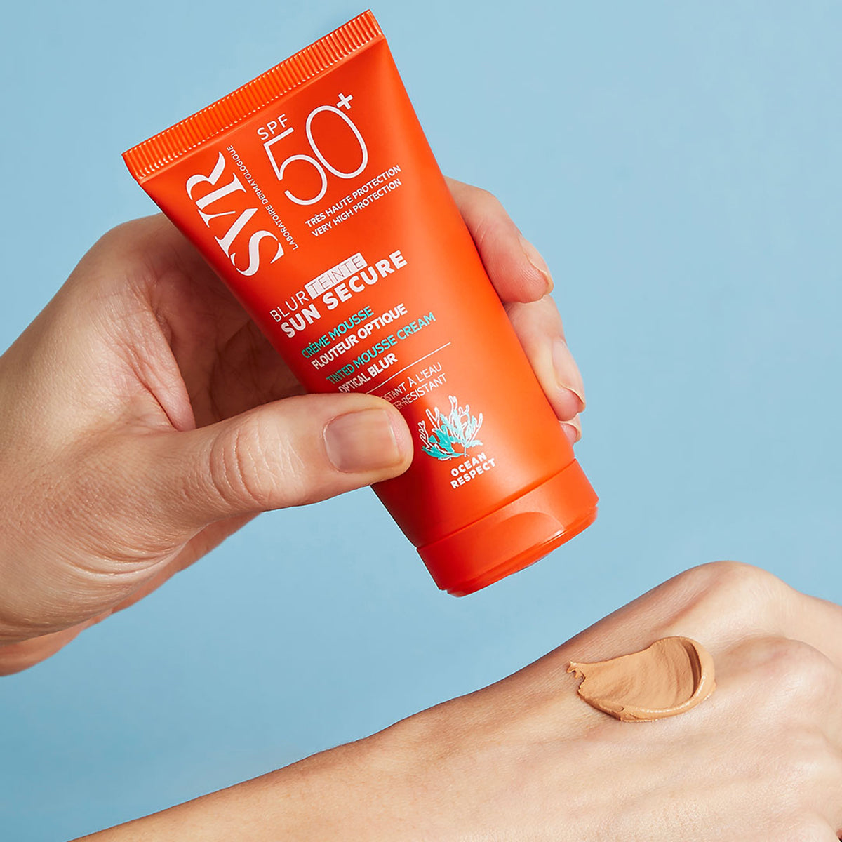 Tubo laranja de SVR Sun Secure Blur SPF50+ com textura em mousse cremosa aplicada na mão sobre fundo azul.