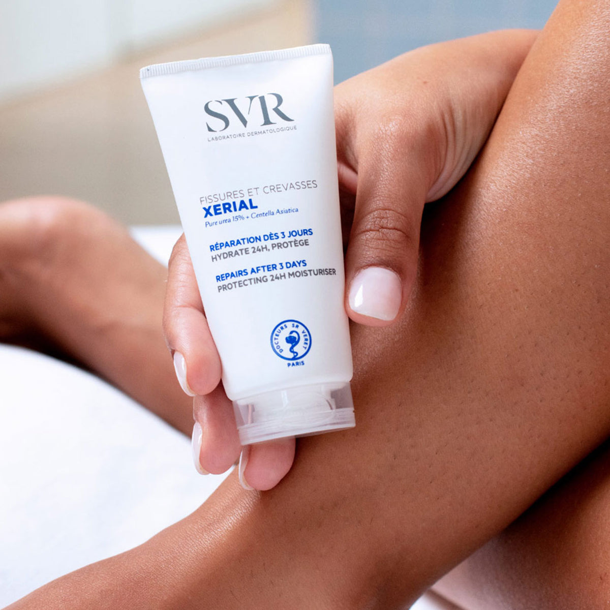 Tubo branco de creme reparador SVR Xerial Fissures & Crevasses sendo segurado por uma mão sobre uma perna hidratada.