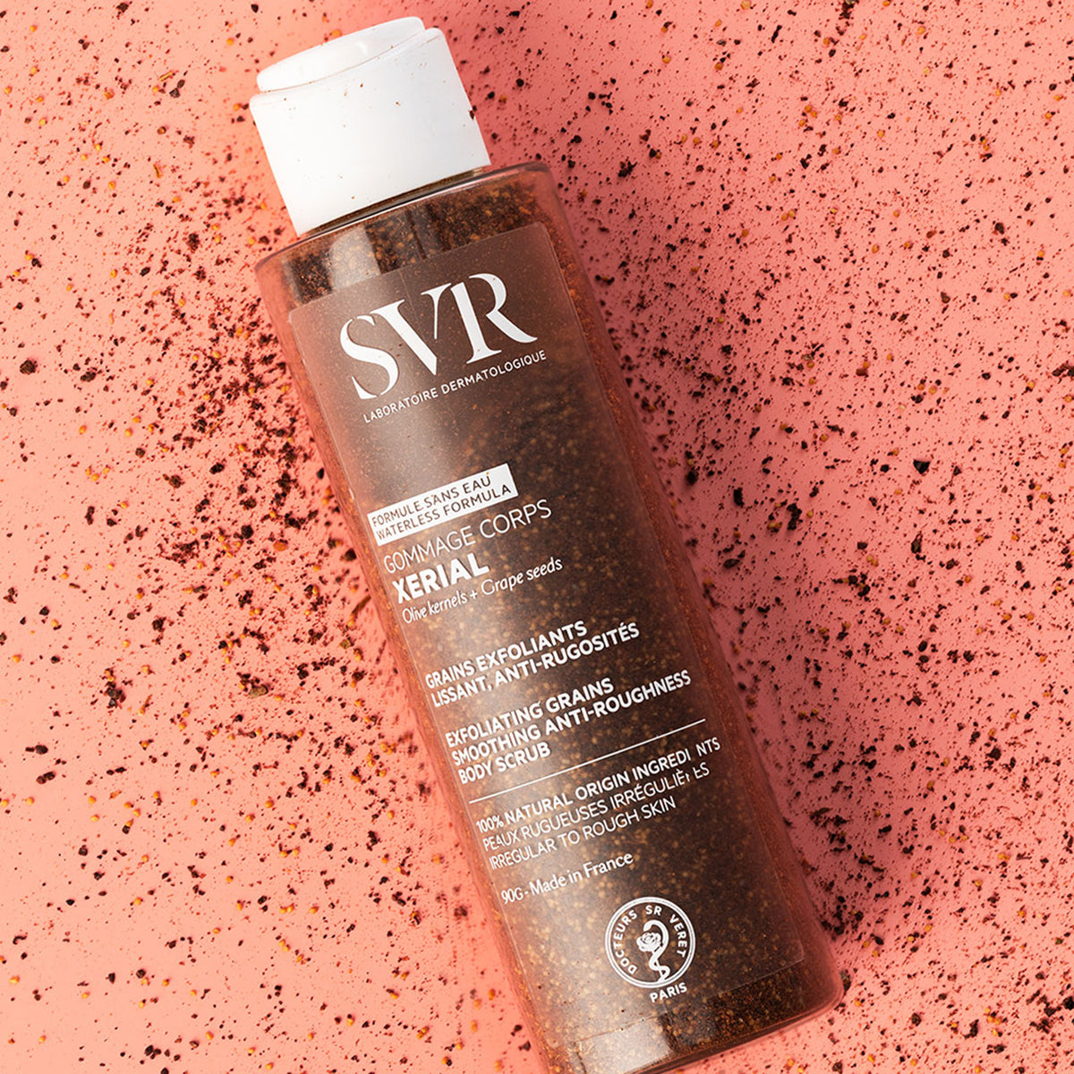 Embalagem cilíndrica transparente do esfoliante SVR Xerial Gommage com grânulos naturais sobre fundo texturizado cor de rosa.