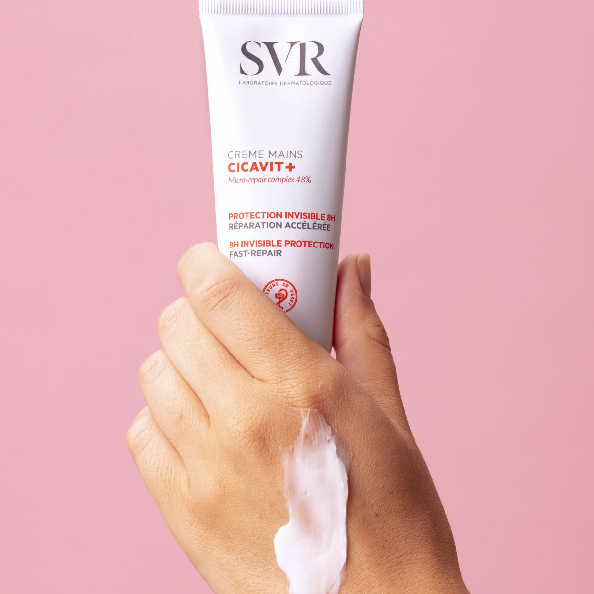 Mão segurando tubo branco de creme de mãos SVR Cicavit+ com textura cremosa aplicada na pele sobre fundo rosa.