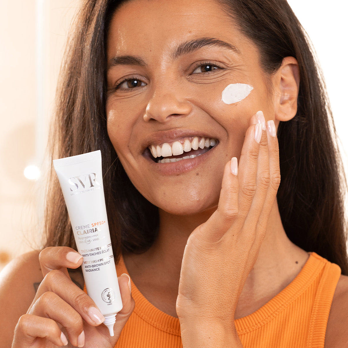 Mulher sorridente aplicando creme facial SVR Clairial SPF50+ branco na bochecha, segurando a bisnaga do produto.