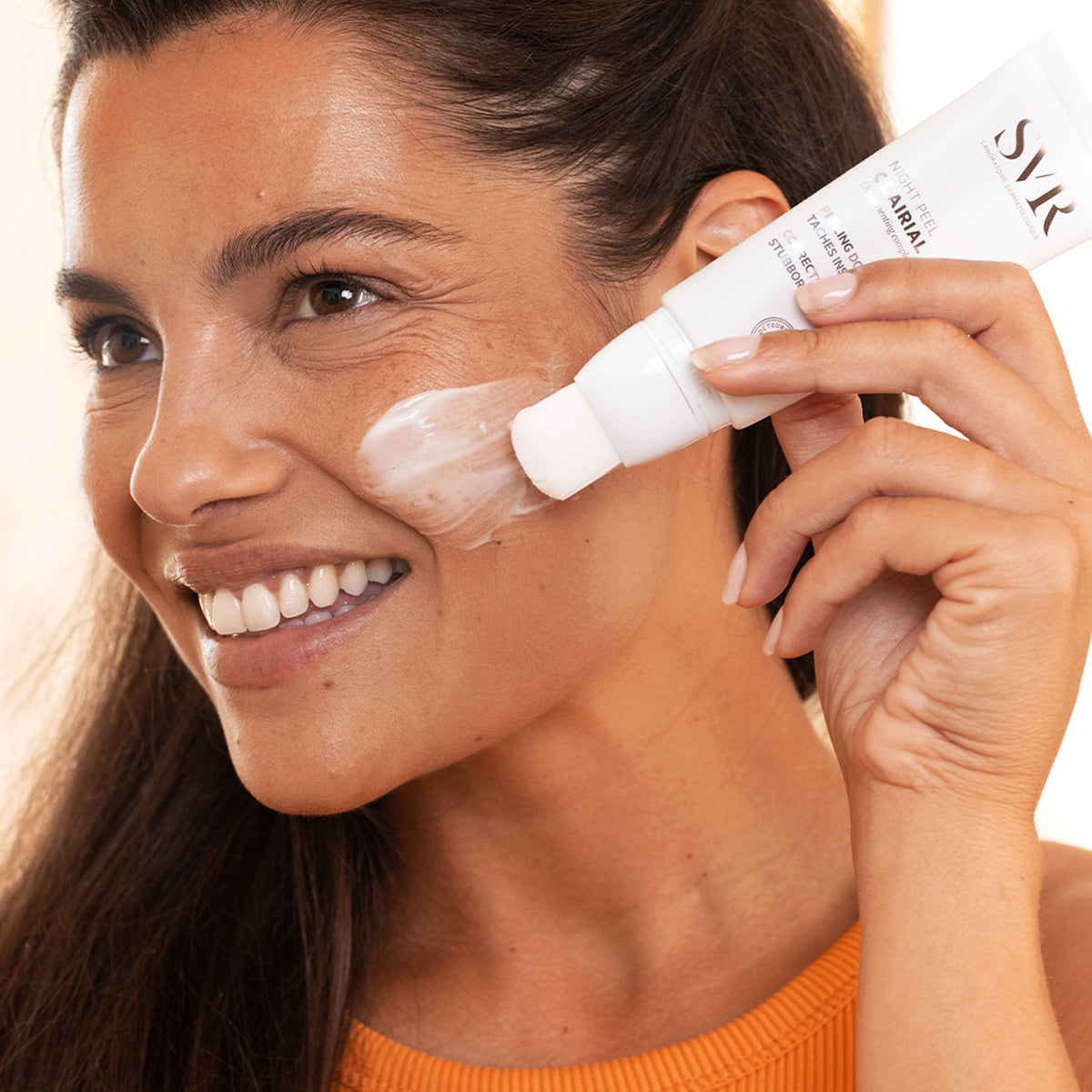 Mulher sorridente aplicando o peeling facial SVR Clairial Night Peel com o aplicador de pincel integrado na bochecha.