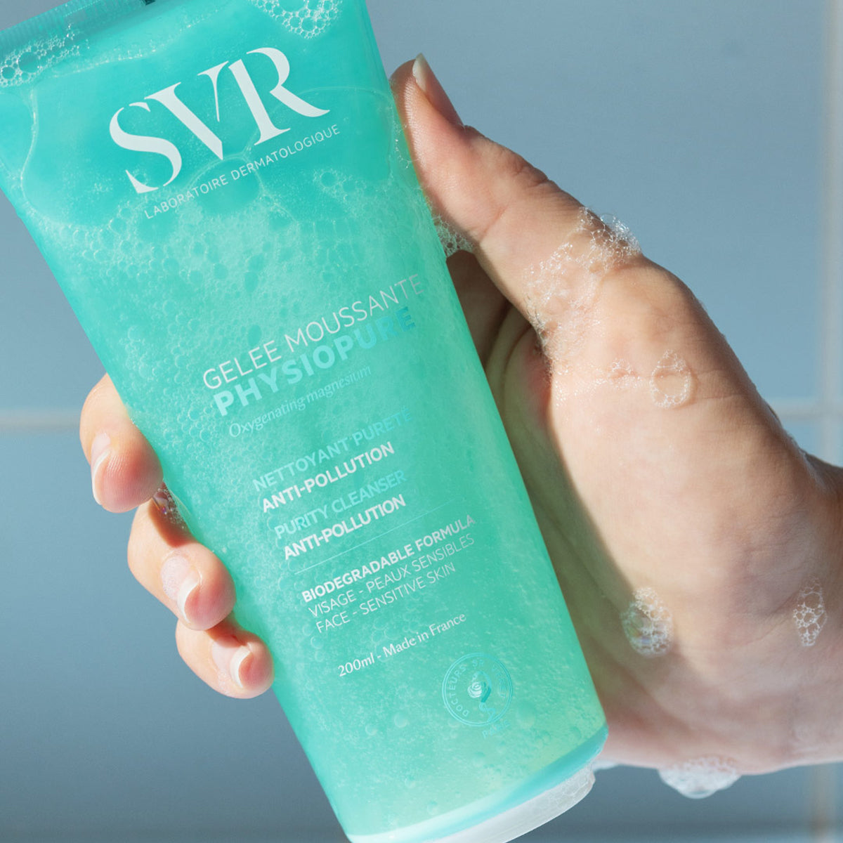 Embalagem de 200ml do gel de limpeza SVR Physiopure em tom azul-turquesa, sendo segurada e criando espuma suave na mão.