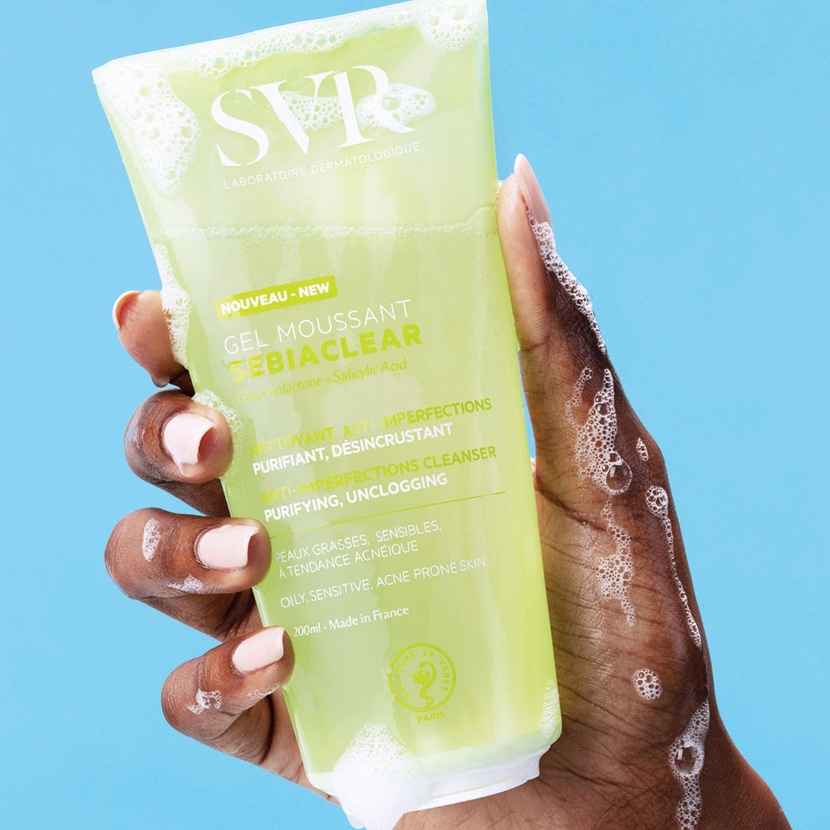 Mão segurando bisnaga verde de 200ml do gel de limpeza SVR Sebiaclear com espuma sobre fundo azul.