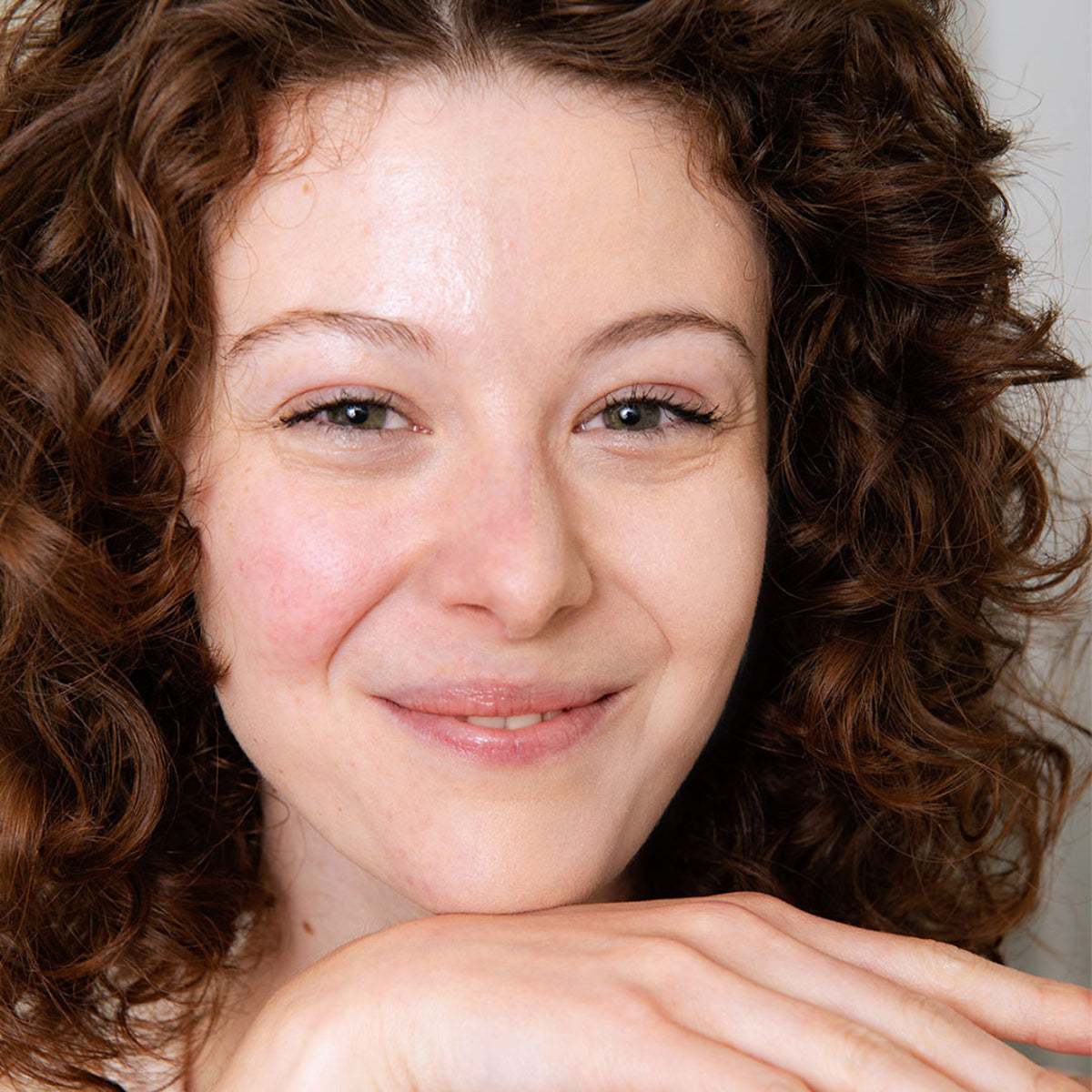 Mulher jovem de pele clara e cabelo castanho encaracolado sorrindo com a mão no queixo, focada em cuidados para pele sensível.