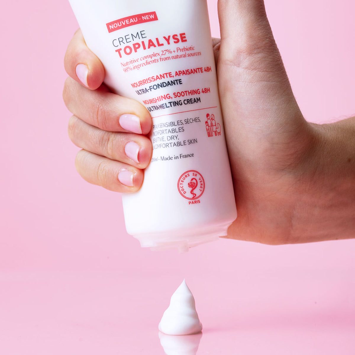 Mão a segurar bisnaga branca de creme TOPIALYSE com textura aveludada a sair sobre fundo rosa suave.
