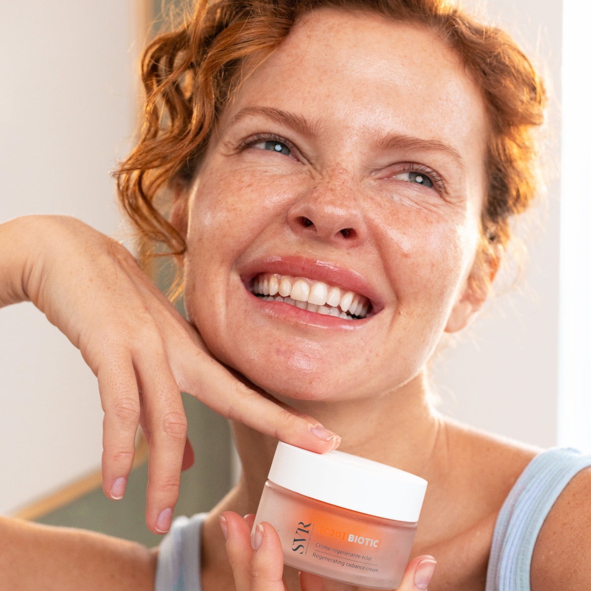 Smiling woman with red hair holding a jar of SVR C20 Biotic regenerating radiance cream in a frosted glass container.