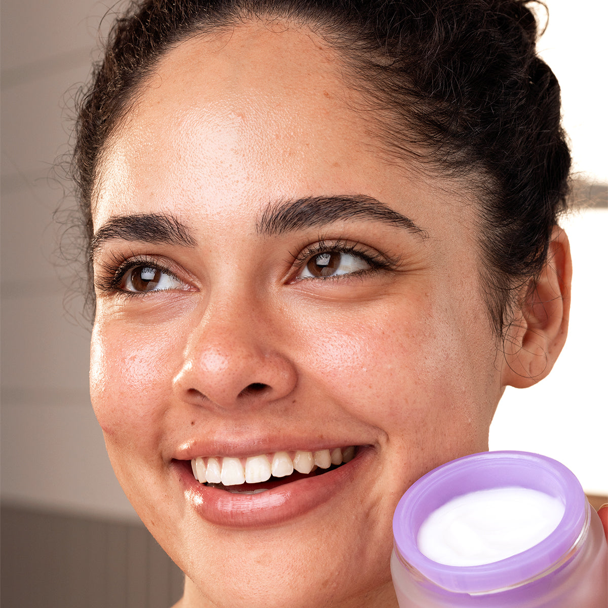 Mulher sorridente com pele radiante segurando um frasco de creme facial PEPTI Biotic com tampa lilás e textura cremosa.