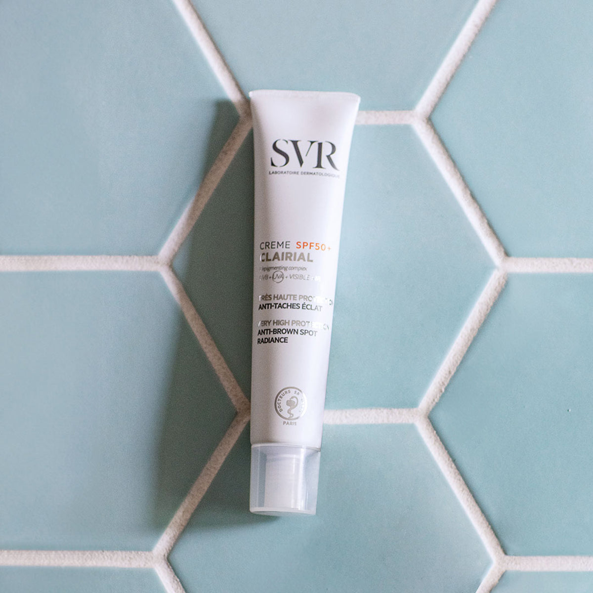 Tubo branco de creme SVR Clairial SPF50+ sobre um fundo de azulejos hexagonais em tom azul-claro.