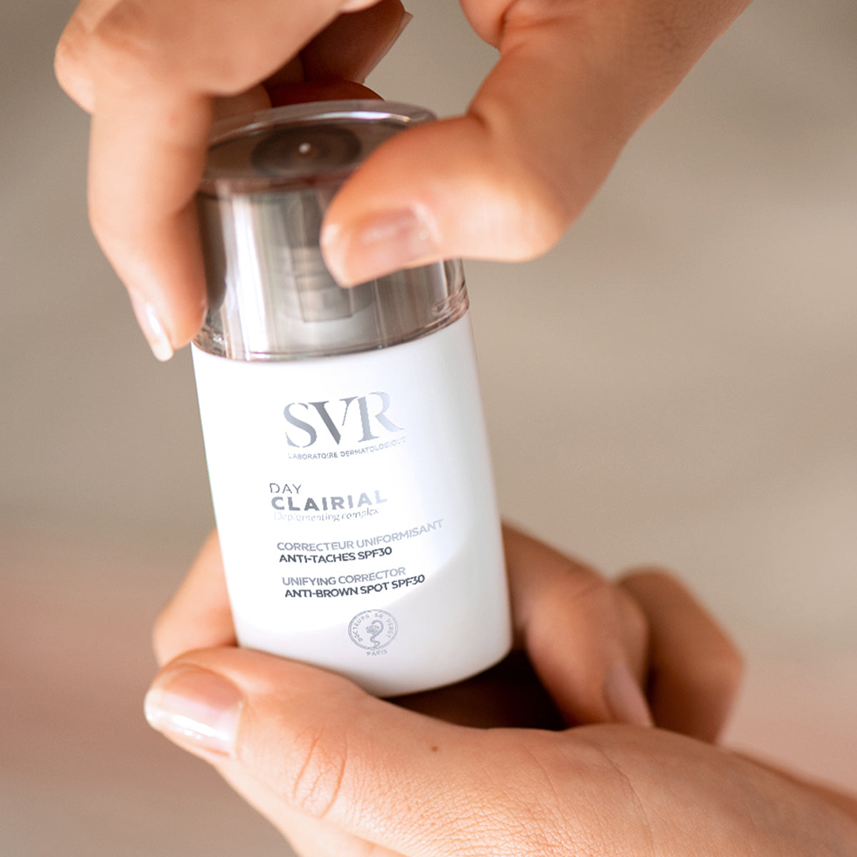 Frasco branco de SVR Clairial Day com tampa transparente sendo segurado por mãos, creme antimanchas com proteção SPF30.