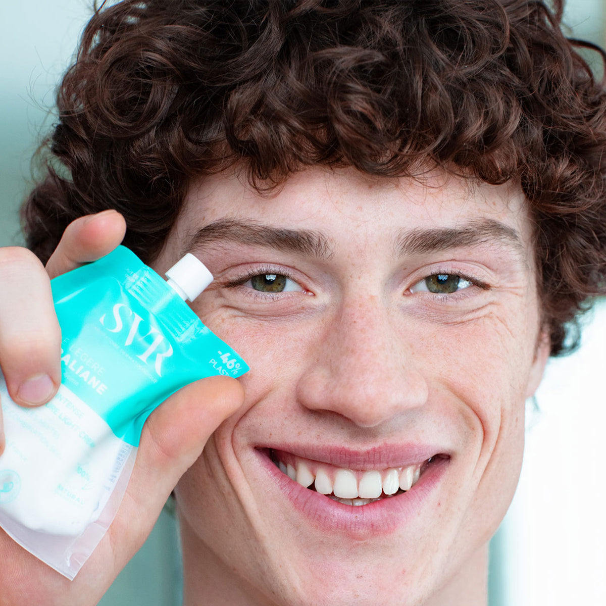 Jovem sorridente segura embalagem flexível e sustentável do creme hidratante SVR Hydraliane Legere em tom azul-turquesa.