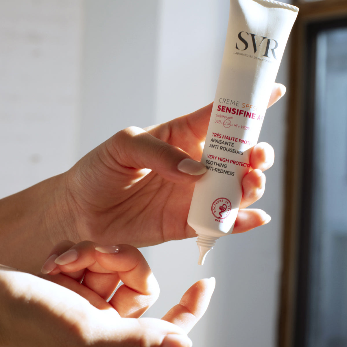 Tubo branco de creme SVR Sensifine AR SPF50+ sendo segurado por mãos, com foco na embalagem anti-vermelhidão.