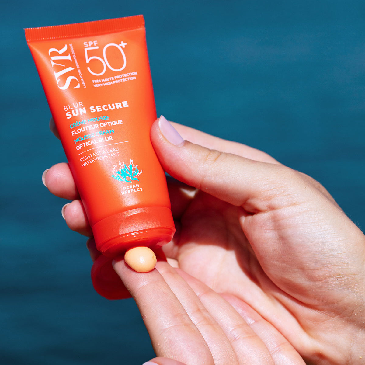 Tubo laranja de protetor solar SVR Sun Secure Blur SPF50+ com textura mousse sendo aplicado na mão sobre fundo azul.