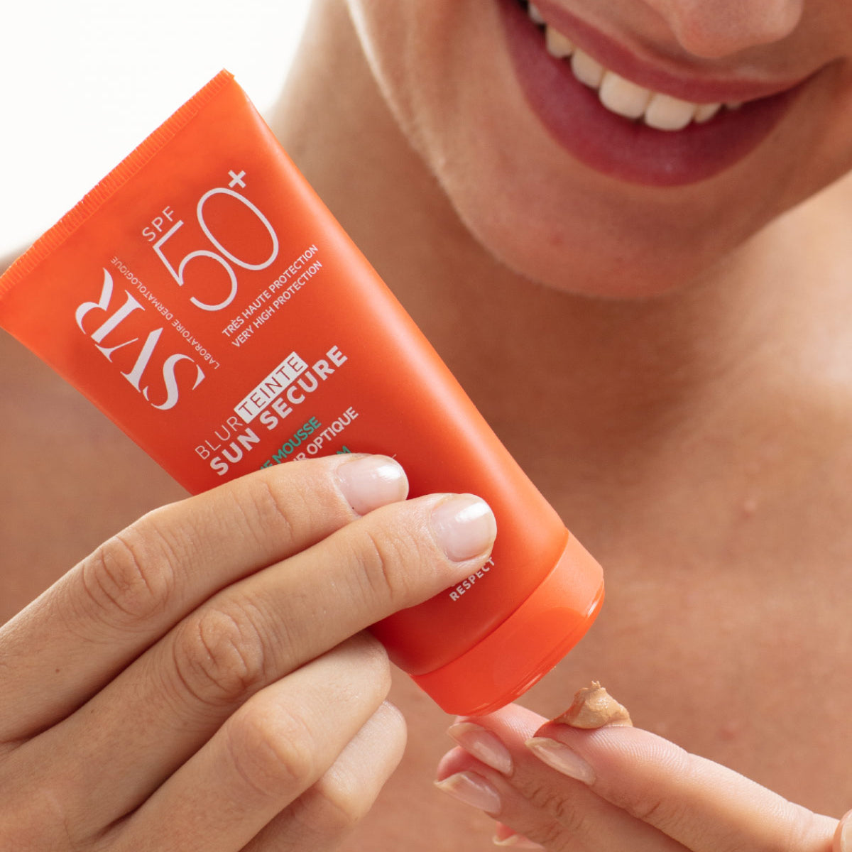 Tubo laranja de SVR Sun Secure Blur SPF50+ com textura em mousse aplicada na ponta do dedo de uma mulher sorridente.