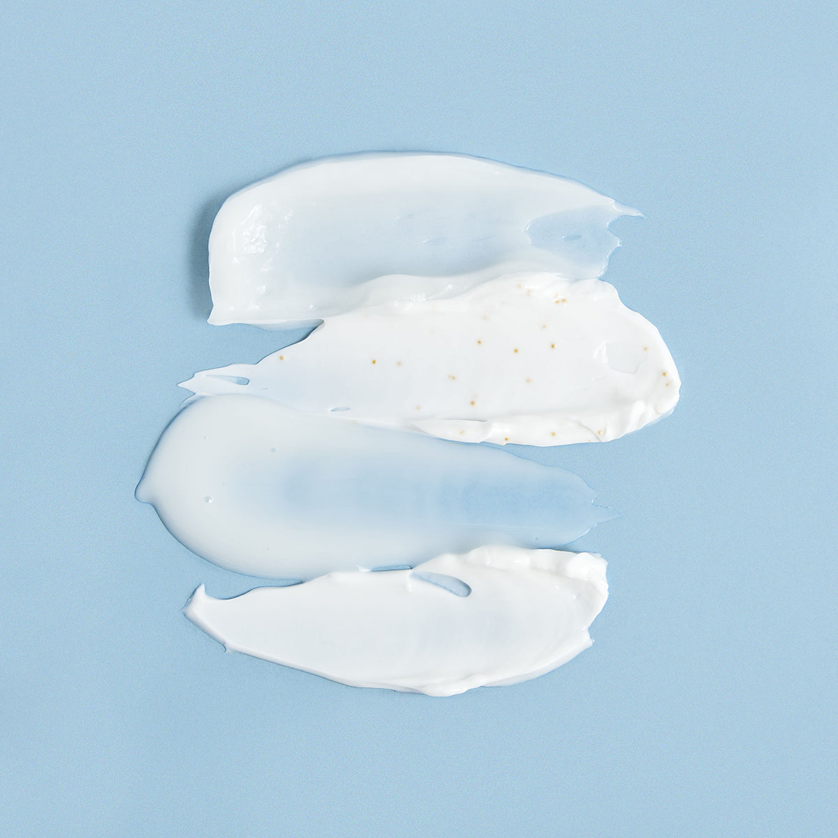 Amostras de creme facial branco com textura suave e partículas douradas sobre um fundo azul claro.