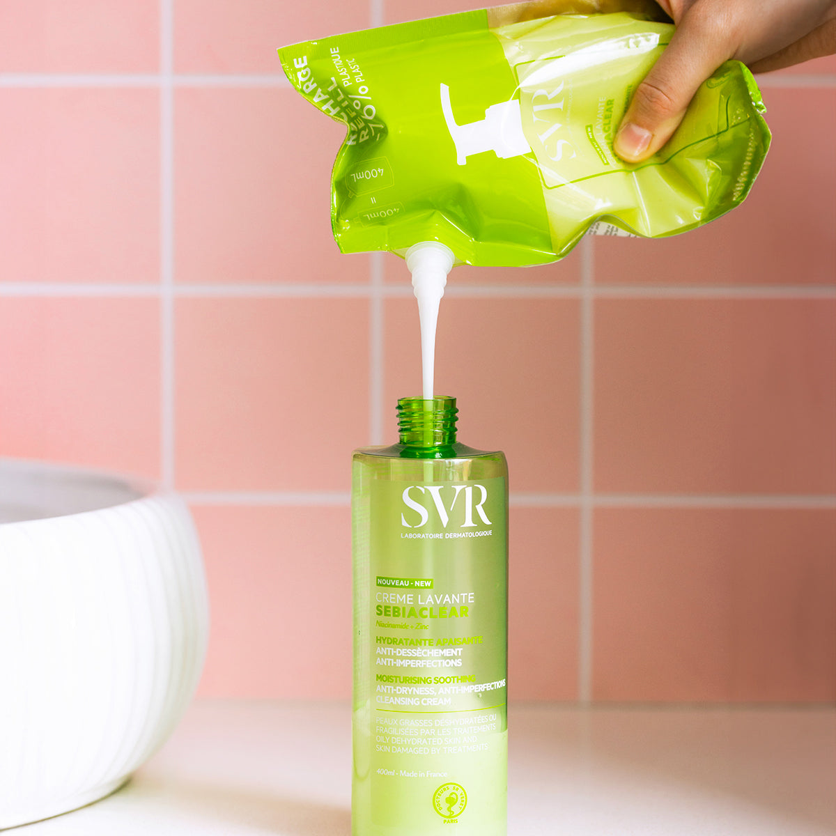 Recarga de SVR Sebiaclear Creme Lavante a ser vertida numa embalagem verde de 400ml sobre um fundo de azulejos rosa.
