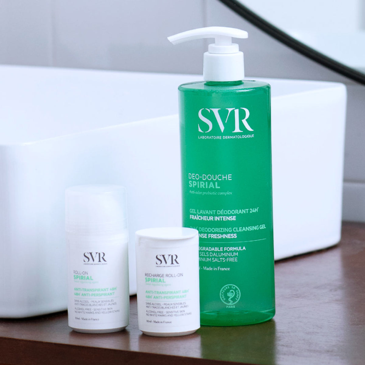 Frasco de gel de duche verde SVR Spirial com doseador e dois desodorizantes roll-on brancos sobre uma bancada de casa de banho.