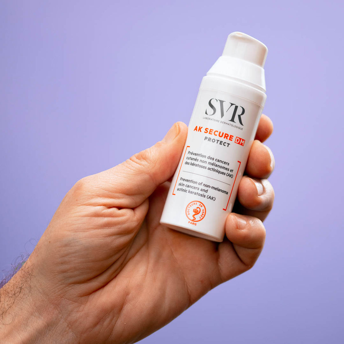 Mão segurando frasco branco de SVR AK SECURE DM Protect com dispensador contra fundo lilás.