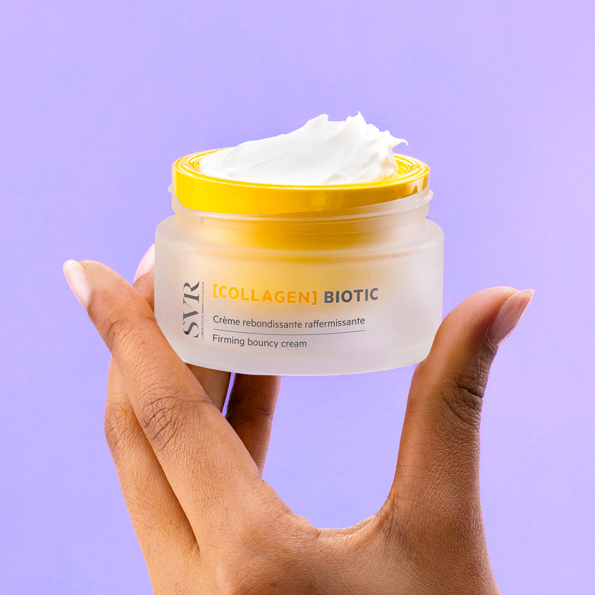 Hand holding a jar of SVR Collagen Biotic firming cream with a bright yellow lid against a light purple background.
