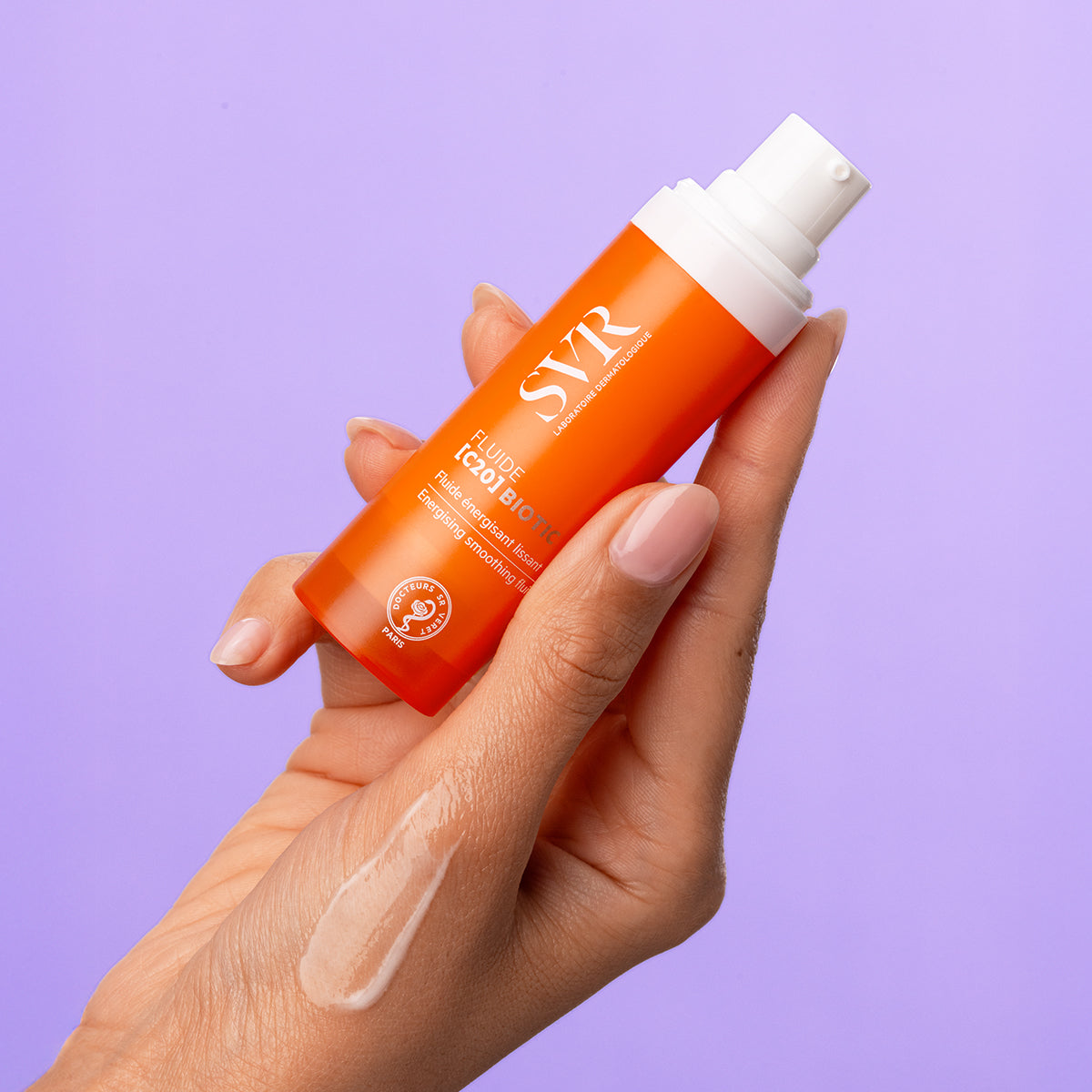 Hand holding an orange SVR Fluide [C20] Biotic pump bottle with a drop of serum on the skin against a purple background.