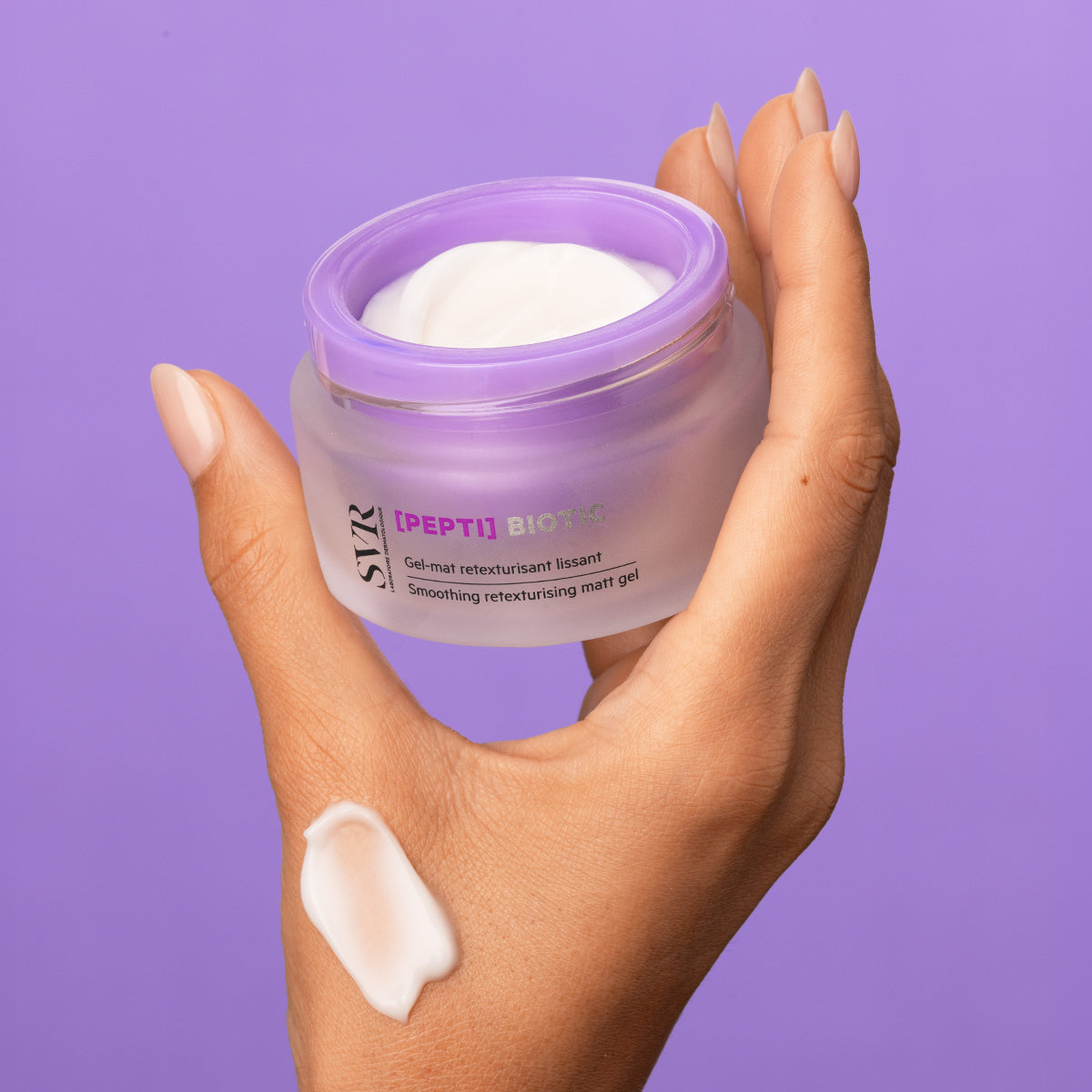 Mão segurando frasco de vidro fosco com tampa roxa do gel regenerador SVR [PEPTI] Biotic e uma amostra de creme branco.