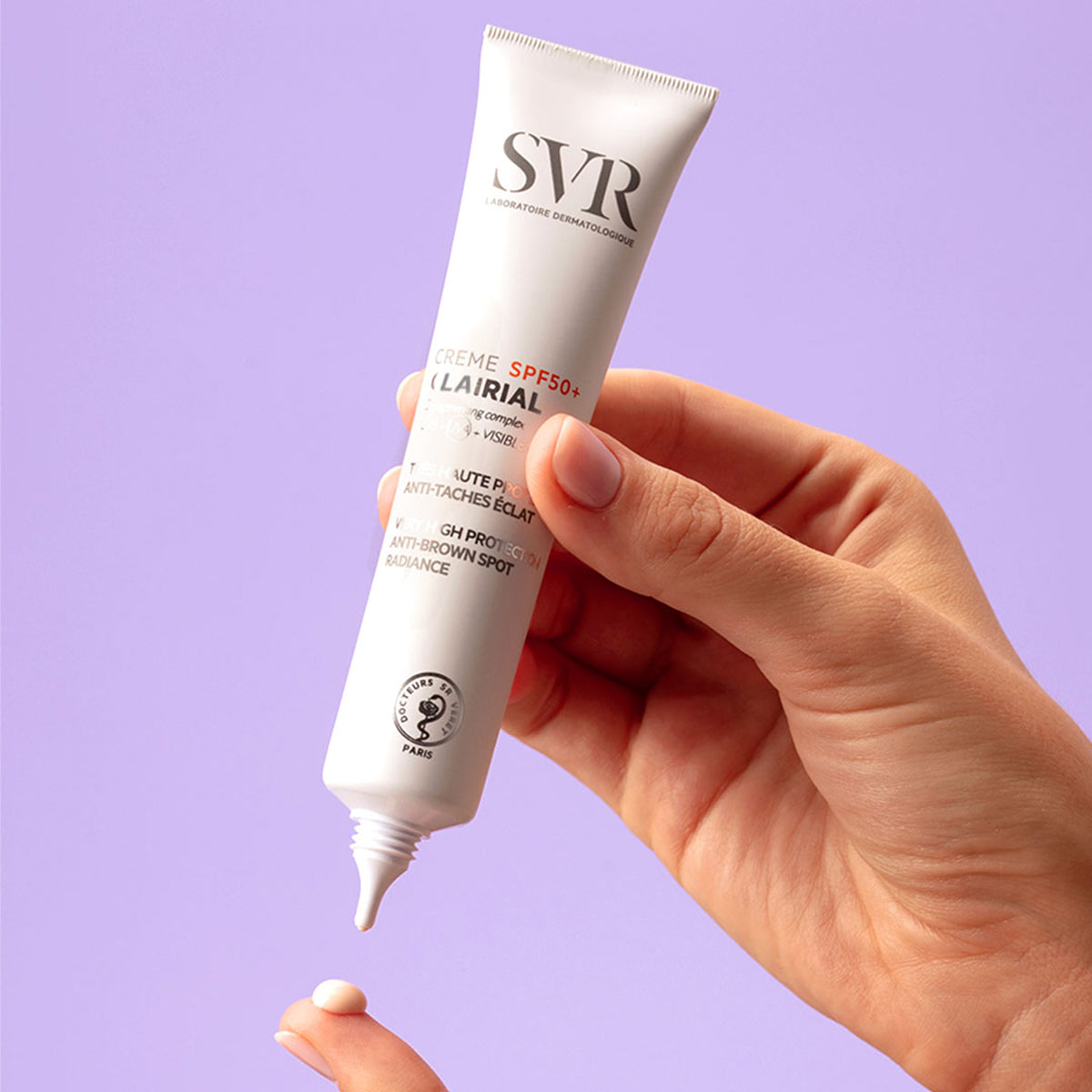 Mão segurando bisnaga branca de SVR Clairial Creme SPF50+ aplicando uma gota de creme na ponta do dedo sobre fundo lilás.