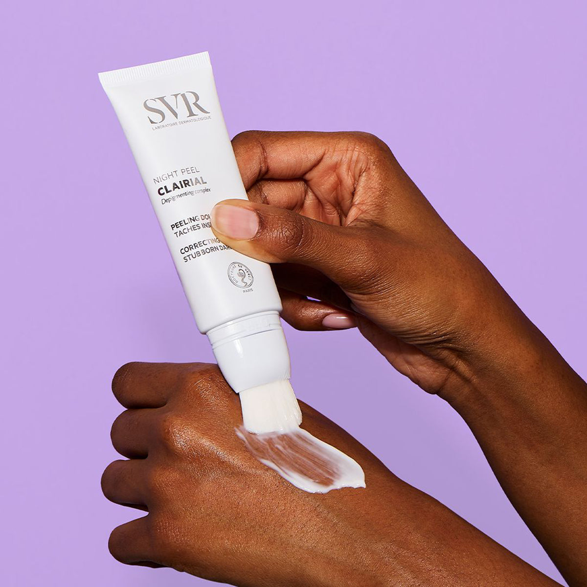 Tubo branco de SVR Clairial Night Peel com aplicador de pincel a aplicar creme na mão sobre fundo lilás.