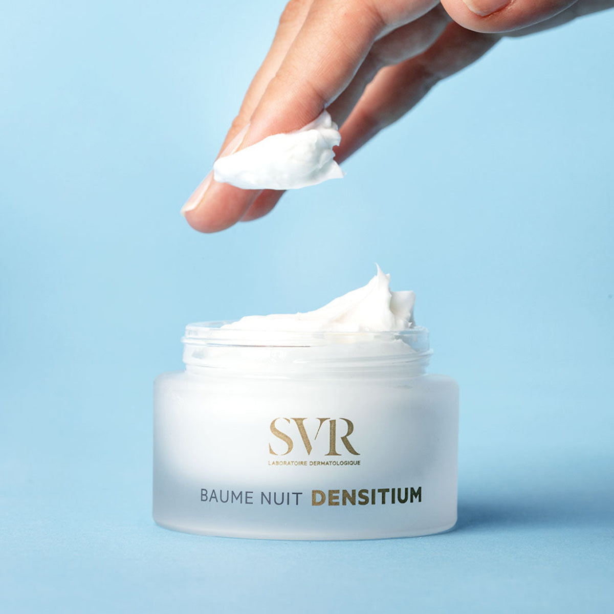 Frasco de vidro fosco do creme SVR Densitium Baume Nuit com textura rica e branca sendo aplicada sobre fundo azul.
