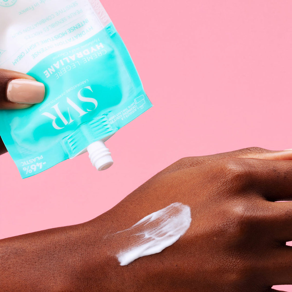 Embalagem azul turquesa do creme SVR Hydraliane Legere sendo aplicada sobre a mão, com textura leve e fundo rosa.