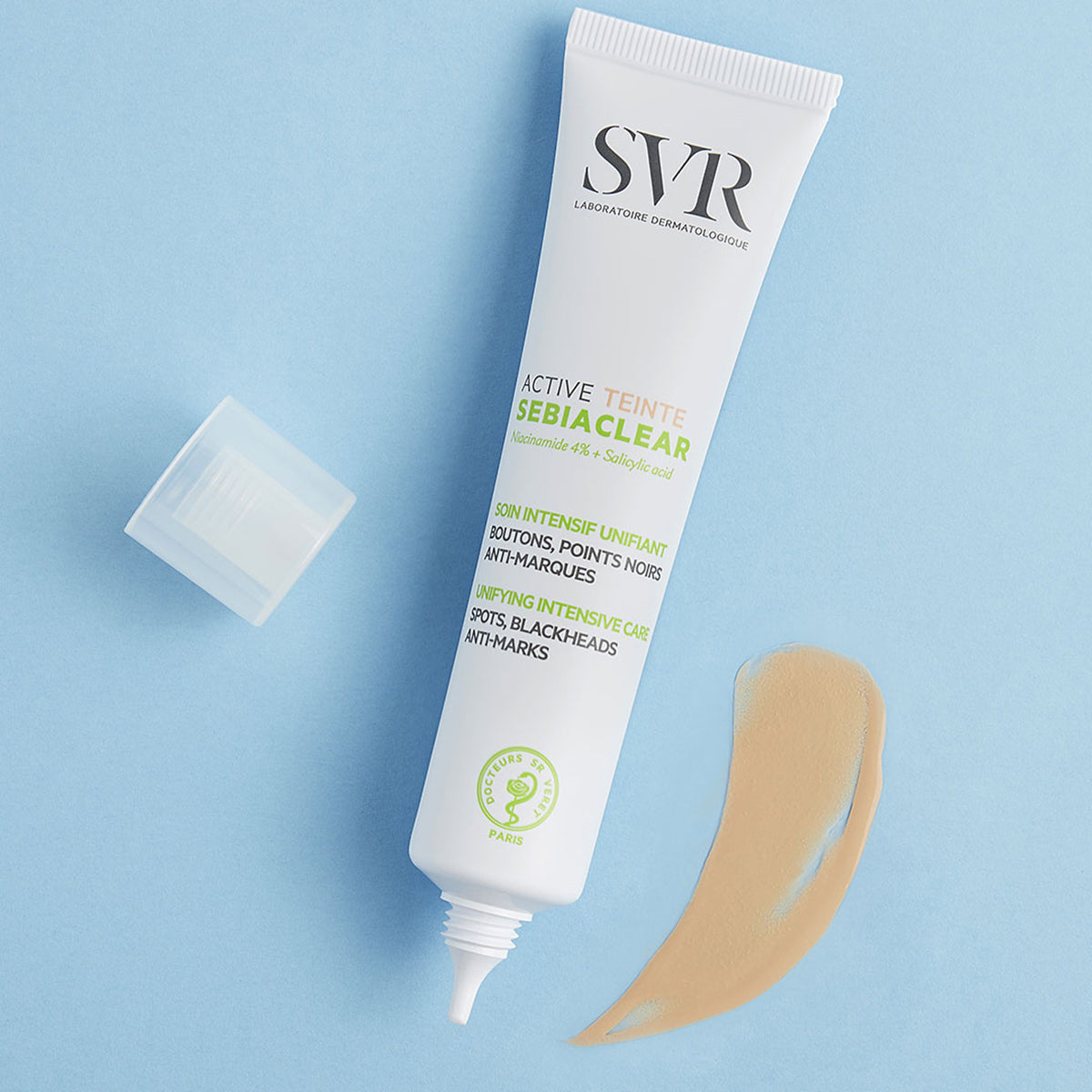 Tubo branco de SVR Sebiaclear Active Teinté com tampa aberta e uma amostra de creme bege sobre fundo azul claro.