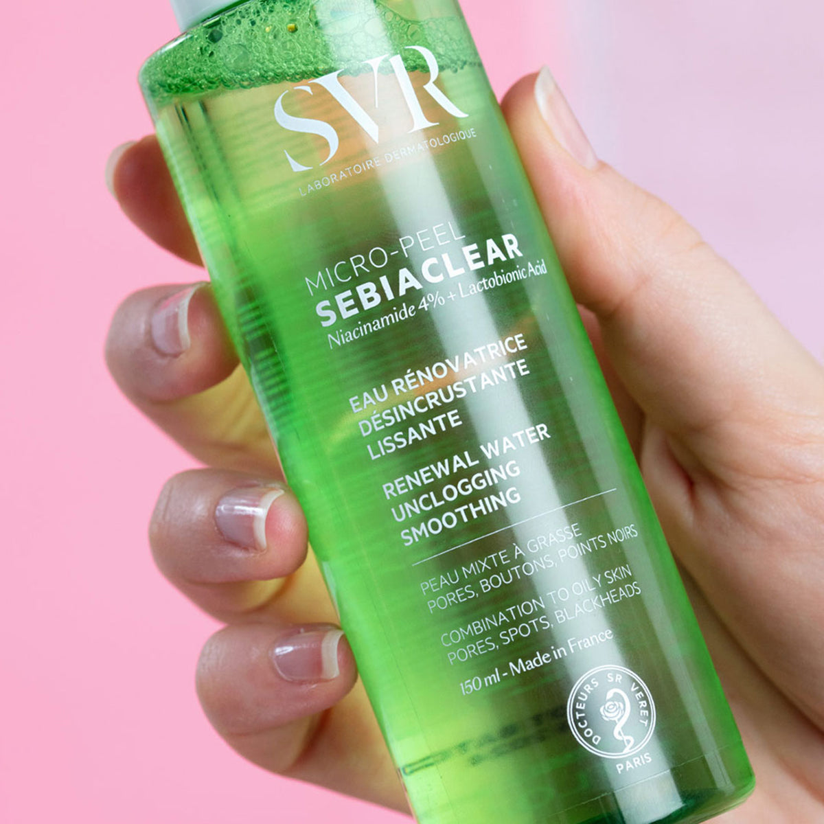 Frasco transparente de 150ml de SVR Sebiaclear Micro-Peel com líquido verde, segurado por uma mão sobre fundo rosa.