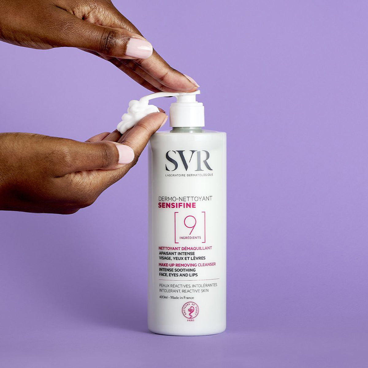Frasco branco de SVR Sensifine Dermo-Nettoyant com doseador, sendo aplicado gel-creme branco numa mão sobre fundo lilás.