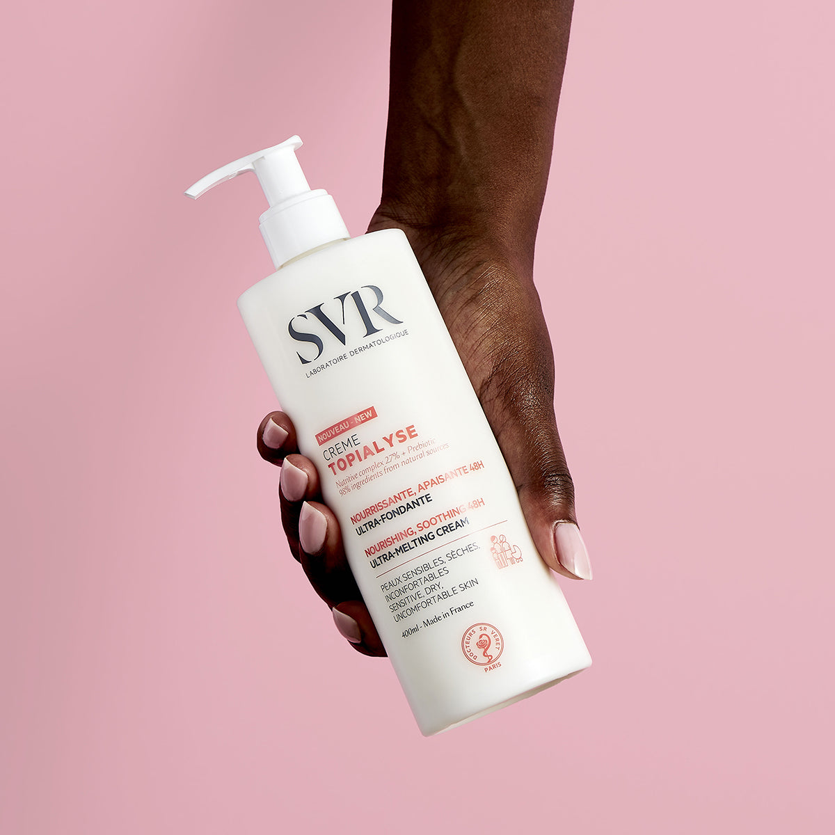 Mão segurando frasco branco de 400ml do creme hidratante SVR Topialyse contra fundo rosa suave.