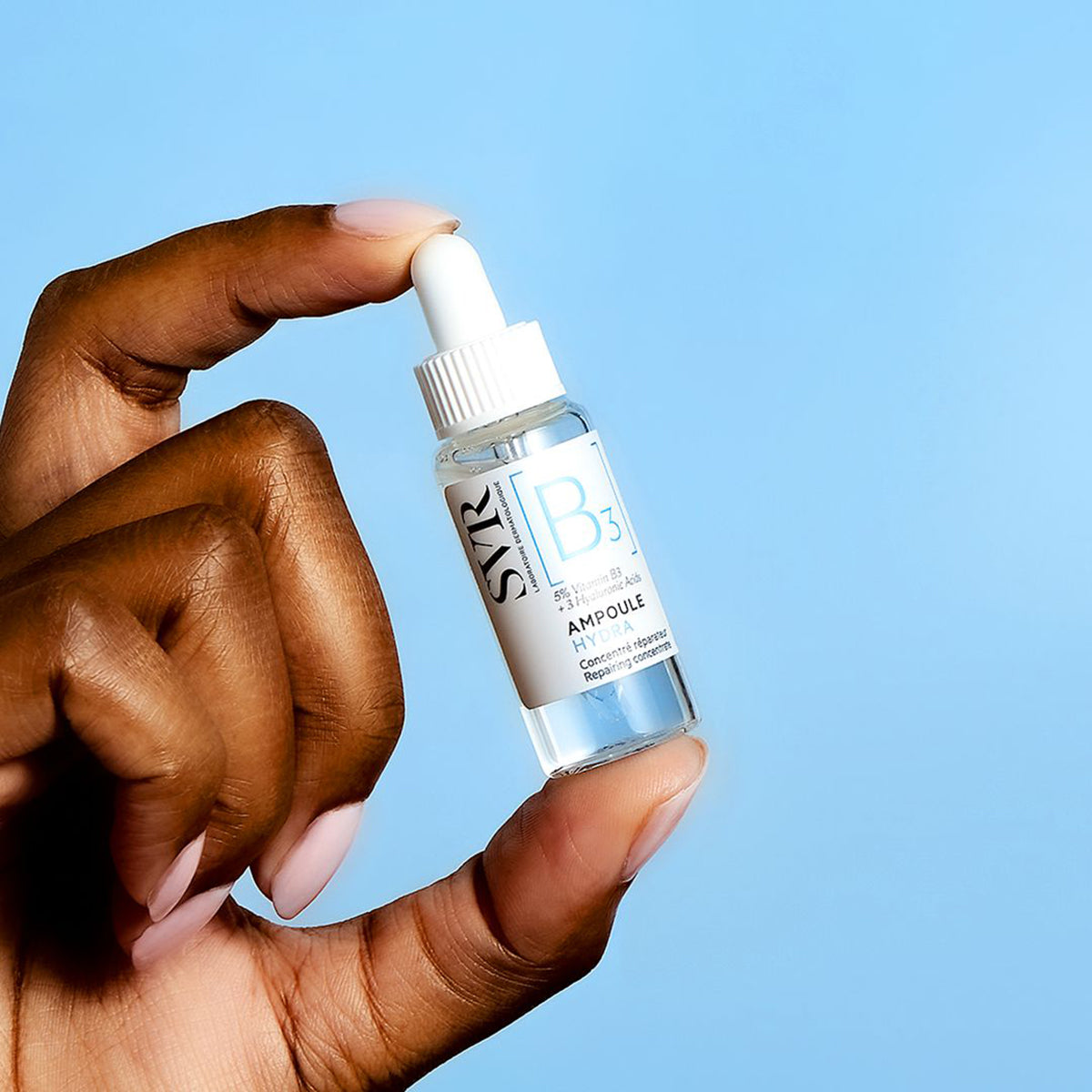 Mão segurando um frasco de vidro transparente SVR [B3] Ampoule Hydra com conta-gotas branco sobre fundo azul claro.