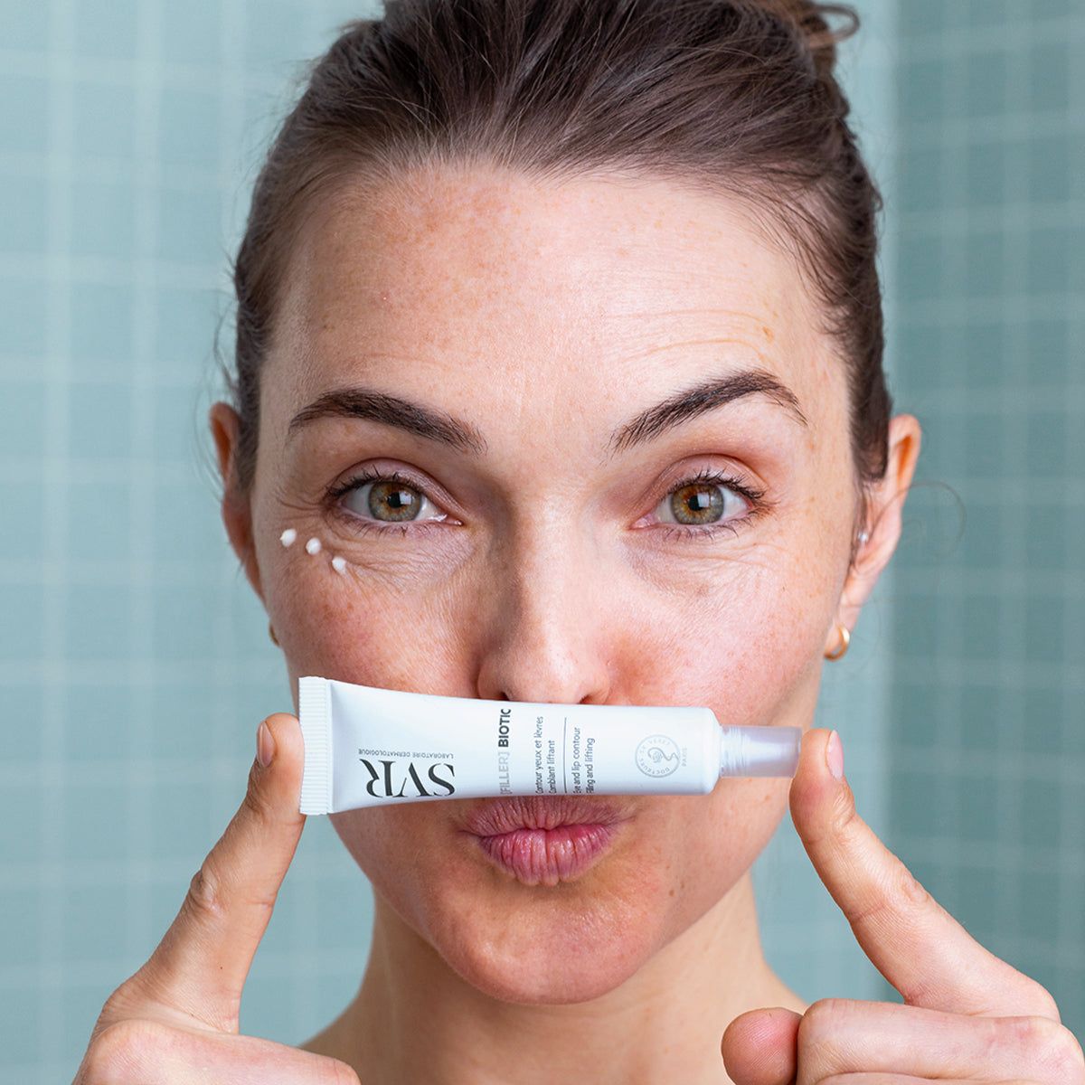 Mulher segura bisnaga branca de creme SVR [FILLER] BIOTIC sob o nariz com pontos de creme no contorno dos olhos.