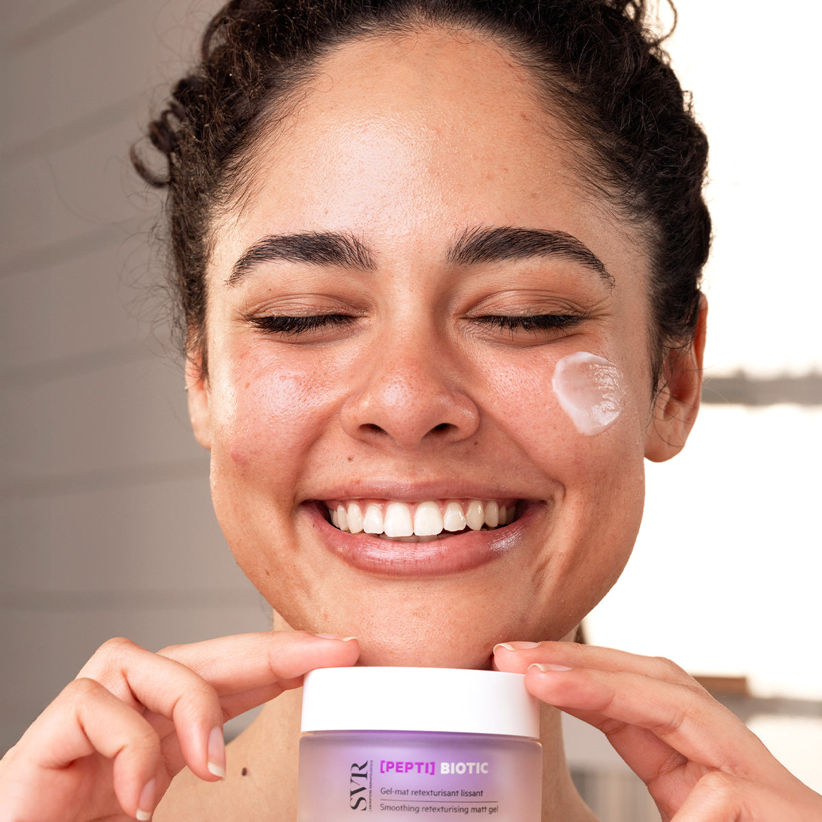 Mulher sorridente com creme SVR [PEPTI] Biotic no rosto, frasco roxo fosco com tampa branca na mão.