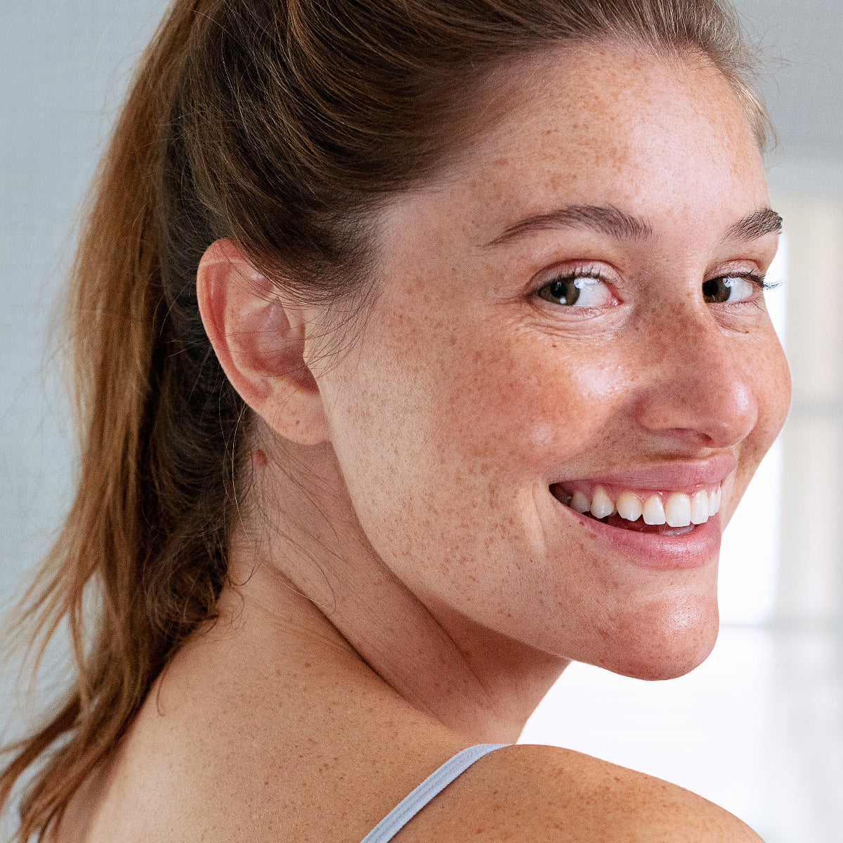 Mulher sorridente com sardas e lábios hidratados e saudáveis, olhando para a câmara com um visual natural e luminoso.