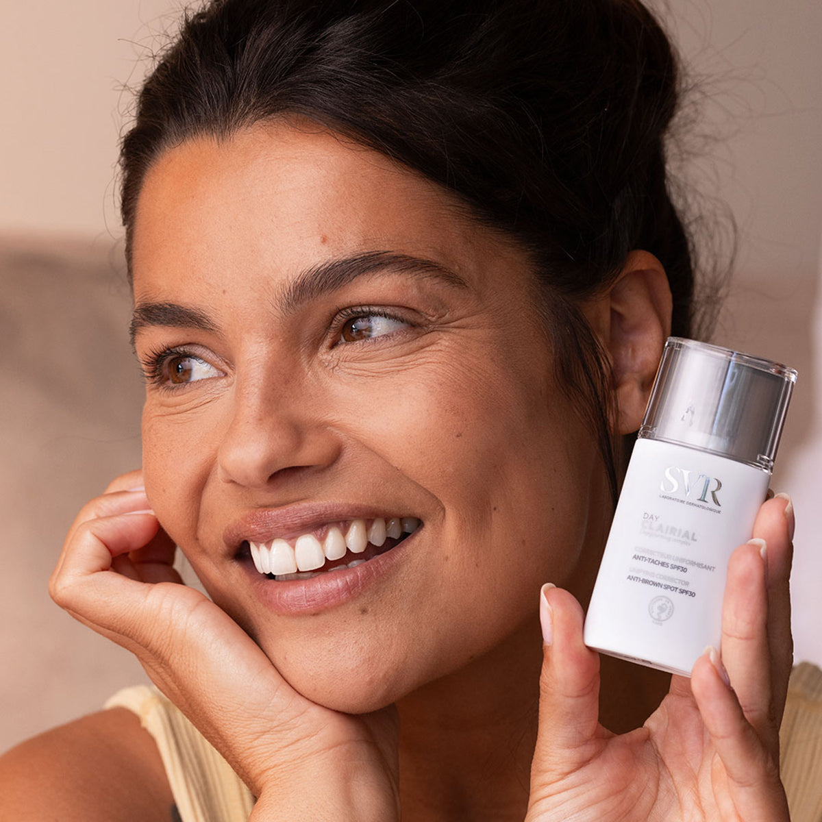Mulher sorridente segura o frasco branco do creme antimanchas SVR Clairial Day com tampa transparente.