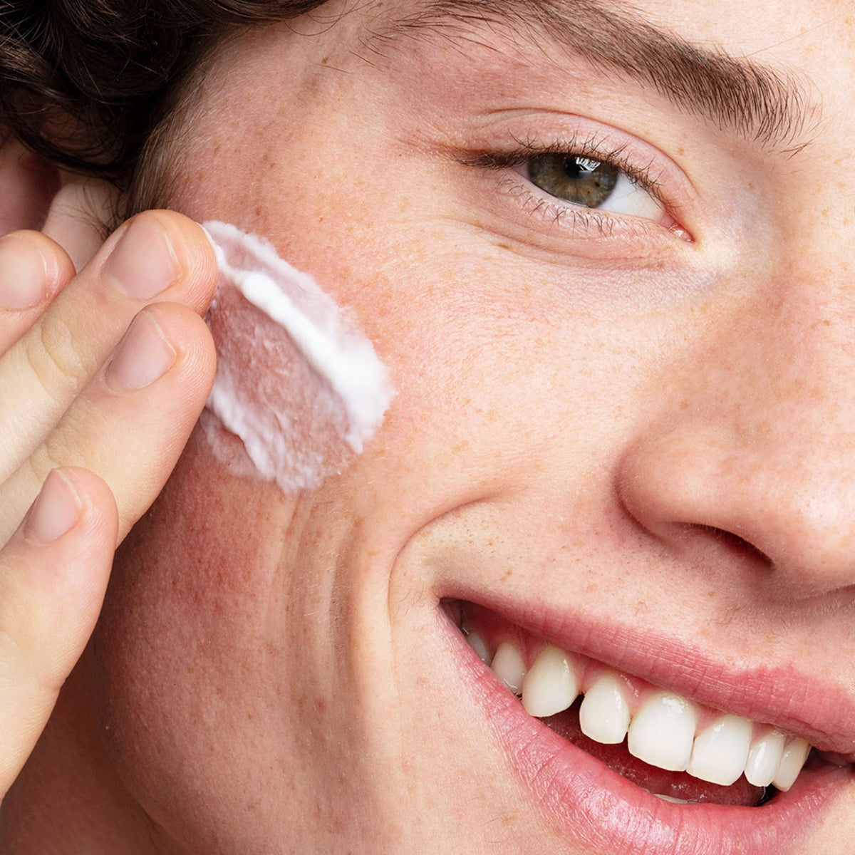 Pessoa sorridente aplicando creme hidratante Hydraliane Legere branco e leve na bochecha, destacando uma pele saudável e natural.