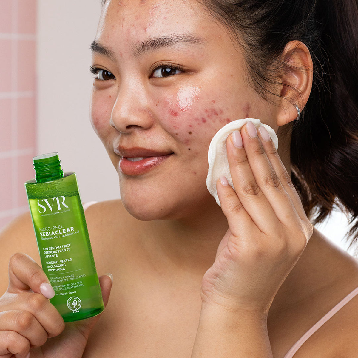 Mulher aplica SVR Sebiaclear Micro-Peel com um disco de algodão no rosto, segurando o frasco verde transparente do produto.