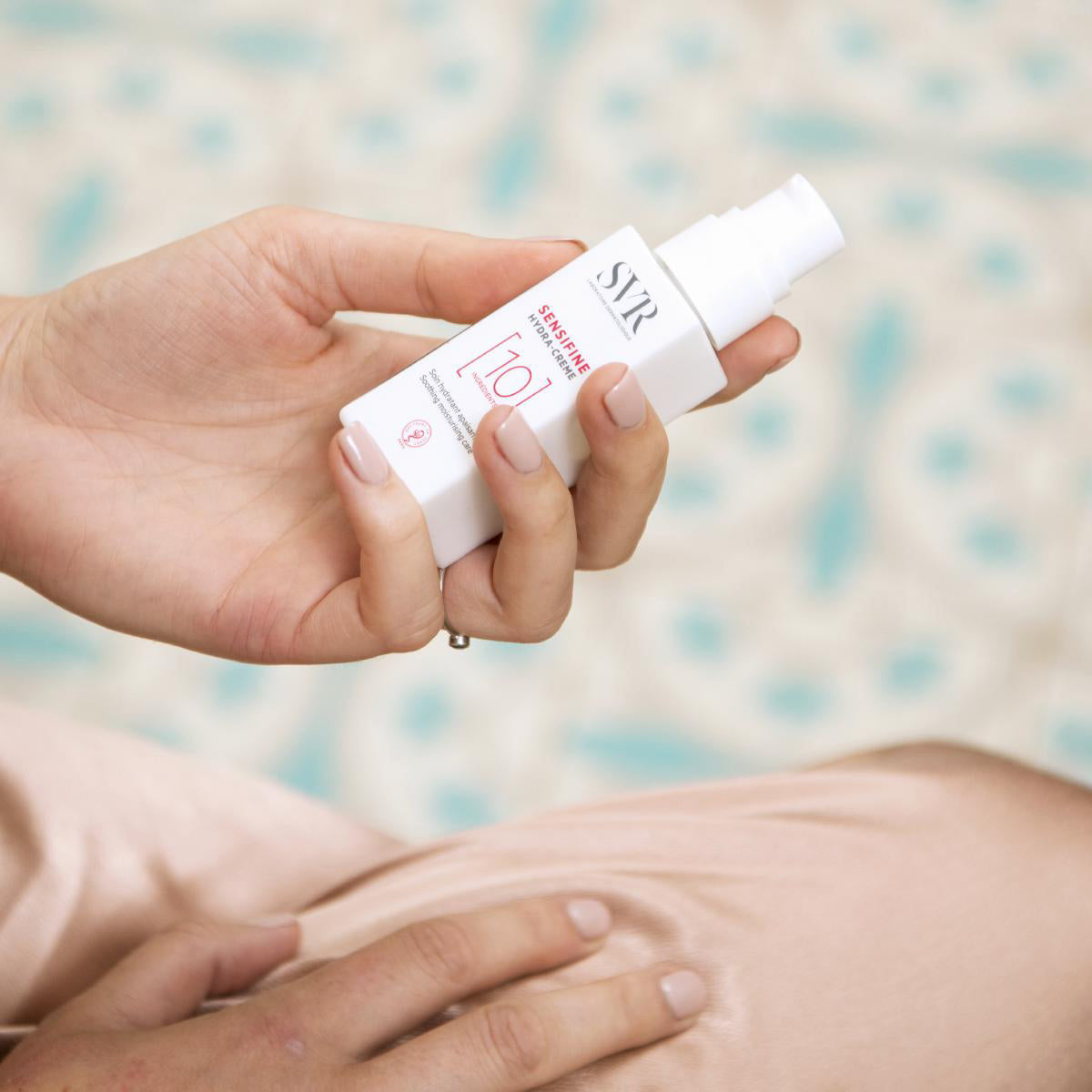 Embalagem branca do creme hidratante SVR Sensifine Hydra-Creme com pump, segurada por uma mão sobre fundo suave.