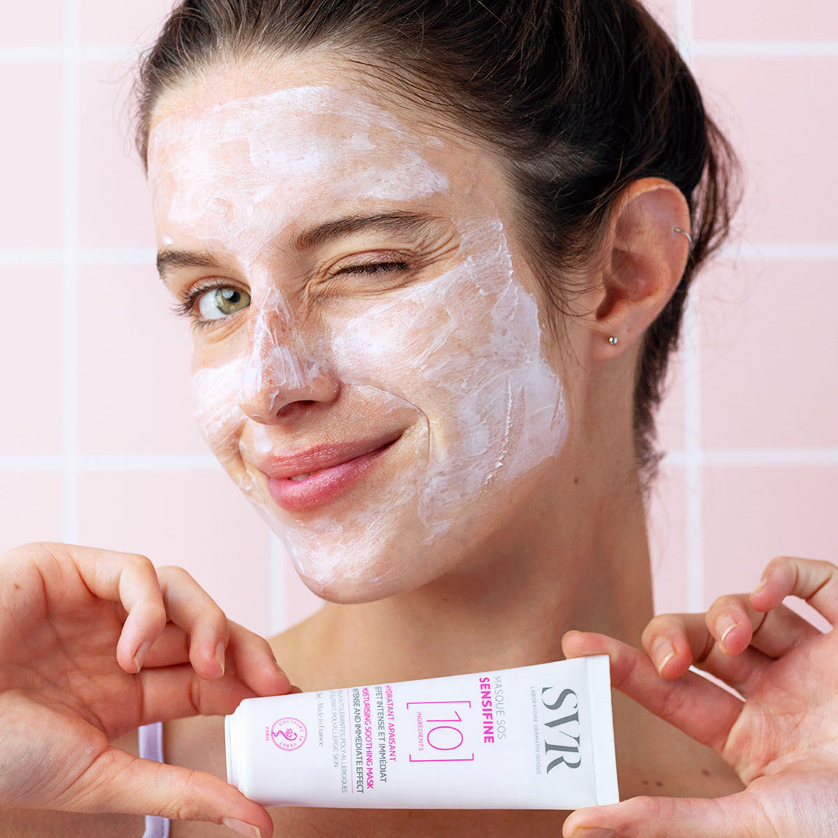 Mulher a aplicar a máscara facial SVR Sensifine SOS branca e cremosa, segurando a embalagem do produto em fundo rosa.