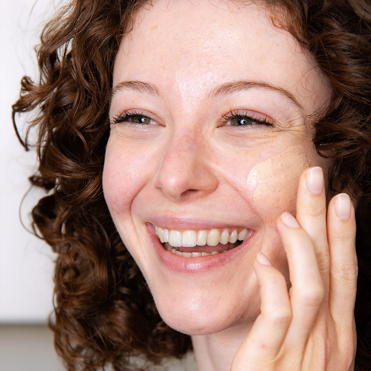 Mulher sorridente aplicando SENSIFINE AR Creme Teintée bege na bochecha para cobrir vermelhidão e uniformizar a tez.