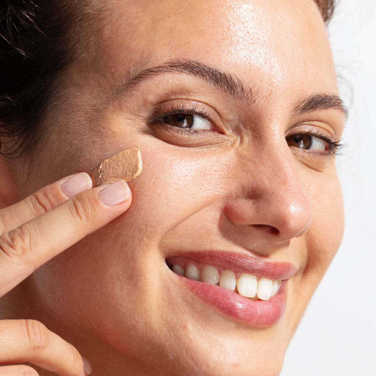Mulher sorridente aplicando creme solar com cor SUN SECURE Blur SPF50+ na bochecha com textura em mousse cremosa.
