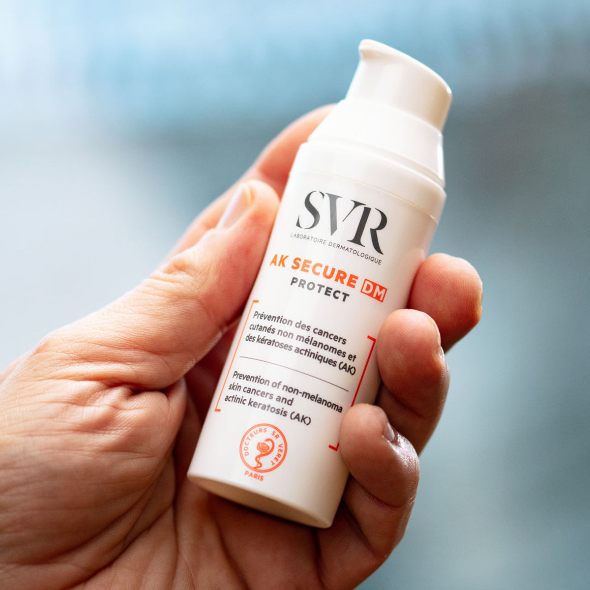 Frasco branco de SVR AK SECURE DM Protect com doseador, segurado por uma mão, exibindo rotulagem informativa em fundo claro.